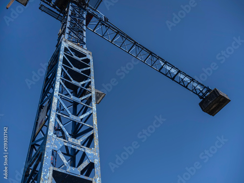 Fotografía A weathered blue tower crane with its counterweight stands against a deep blue sky at a river shipyard