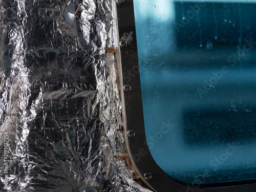 Fotografía Reflective insulation and screwed trim surround the dark frame of a ship's window, blue daylight beyond the glass