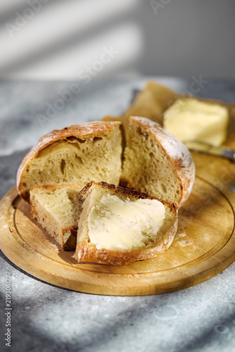 Close-up of freshly baked artisan sourdough bread sliced on a round wooden board. A slice topped with butter in the foreground, rustic food styling.