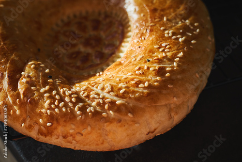 Close-up of golden Uzbek non bread with decorative center pattern and sesame seeds. Traditional Central Asian flatbread on dark background, fresh homemade baking.