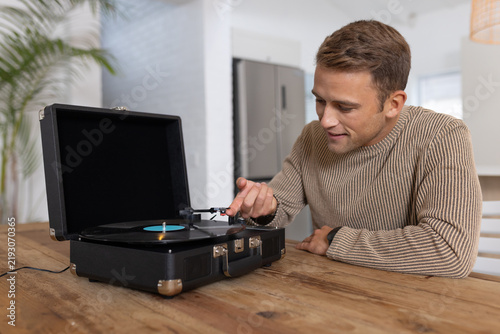 Adult man leaning guiding tonearm toward spinning blue-label vinyl on suitcase turntable in kitchen