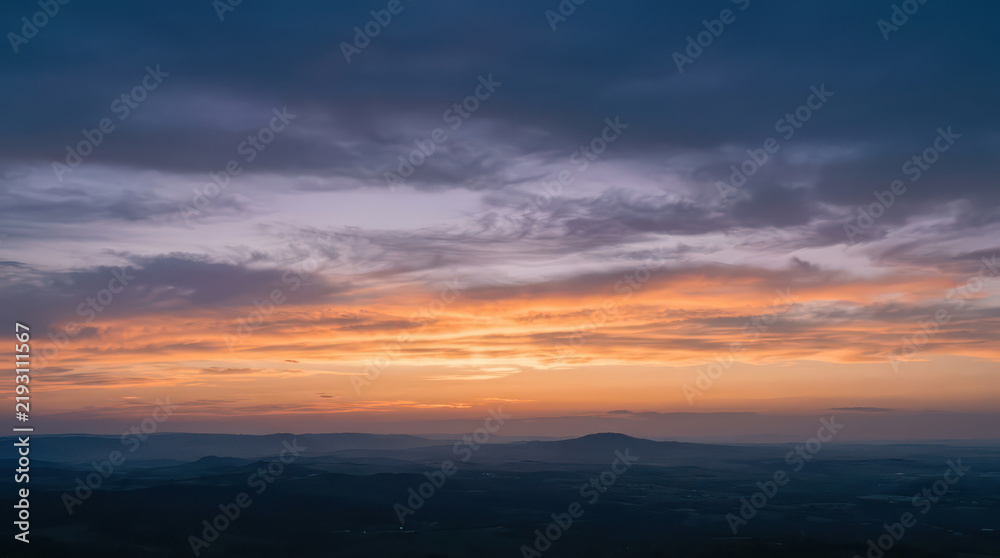 Fototapeta premium Panoramic Sunset Over Layered Hills and Distant Mountain Silhouettes with Vibrant Orange and Blue Sky, Representing Peaceful Landscape, Dusk, Nature Scenery and Golden Hour Atmosphere