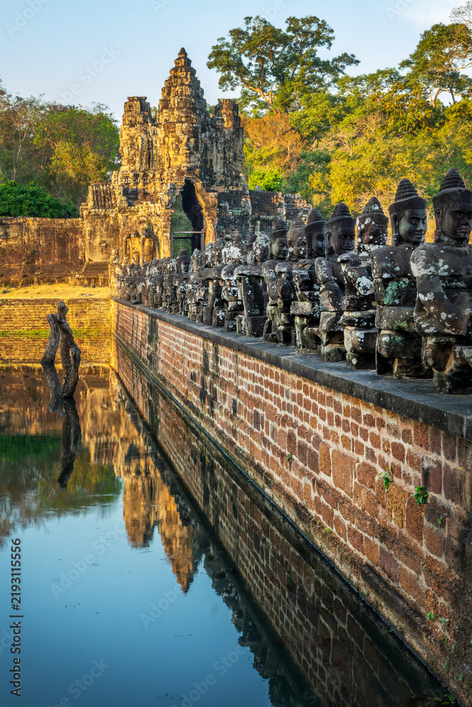 Obraz premium Row of Devas head statues at Angkor Thom South gate, serene Buddha faces at sunset in Angkor, ancient moat with water reflections, Siem Reap, Cambodia
