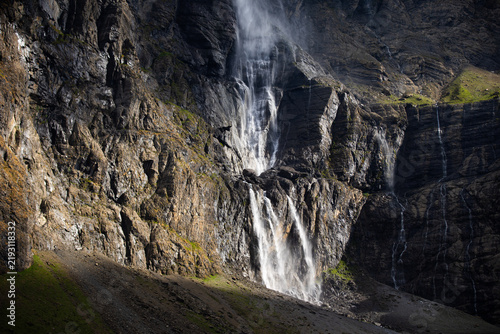 serene waterfall cascading down rocky cliffs in a mountainous landscape from below, looking up the cliff with green and brown vegetation and white rocks creating a dramatic and dynamic view.