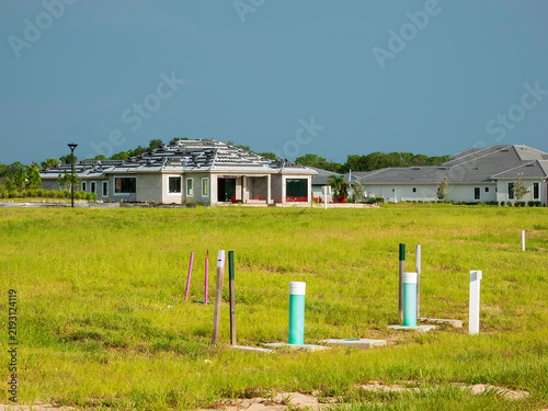 A group of location markers and utility conduits on a buildable lot some distance from a house under construction and a completely roofed house in a suburban residential development in Florida