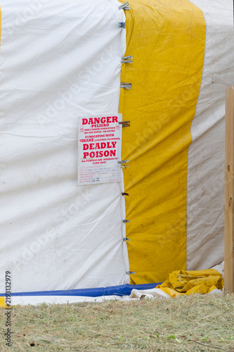 Konstfotografi A yellow-and-white fumigation tent draped over a house behind a wooden fence, showing striped tarpaulin, metal fastenings and a red warning sign indicating poison, restriction and safety precautions