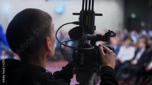 Professional cameraman filming a live conference with video camera. Event broadcasting, live streaming and video production with audience in the background at a business conference.