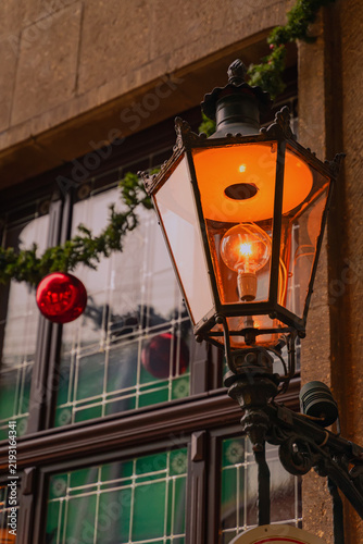 Vintage street lamp glowing beside decorated window. Street lamp glows beside Christmas decorations, shop window in city center. Holiday season, urban architecture, tourism, retail, festive decoration