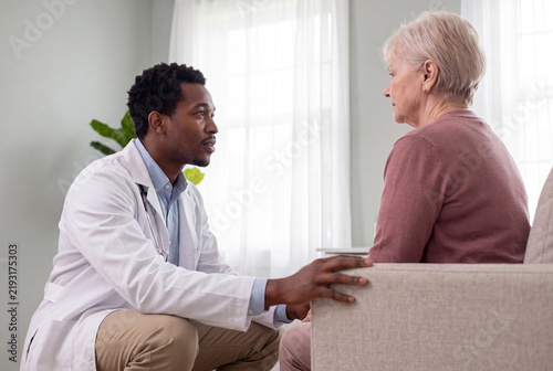 Young African male doctor crouching to a seated senior Caucasian woman patient's eye level to reassure her attentively in a bright calm clinic consultation room

