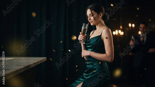 An elegant woman in a green dress enjoys a glass of champagne at a dimly lit bar, with golden confetti falling around her.