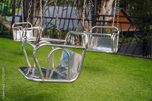 Two shiny metal baby swings hanging outdoors on a vibrant green lawn, framed by a black metal fence and trees, ready for children's playtime in a sunny park setting.