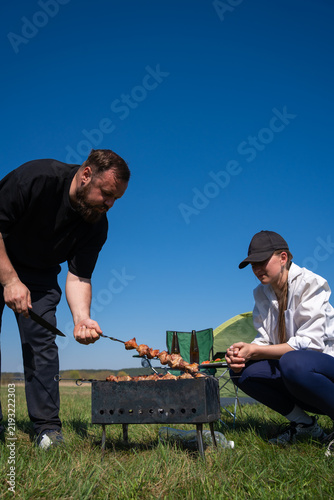 фотографія Man turning skewers with shish kebab on a portable grill while a woman watches, enjoying a relaxed outdoor picnic and camping on a sunny day in a green field with a clear blue sky