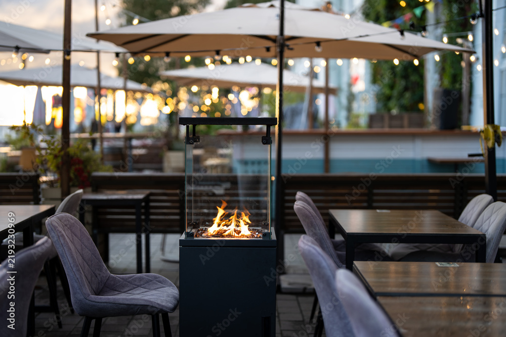 Obraz premium Cozy open-air restaurant terrace with sofas, umbrellas and modern gas fireplace inside glass box, glass patio heater with burning flame beside comfortable dining tables at outdoor cafe settings.