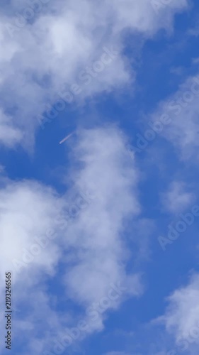 A small rocket flies high through a bright blue sky surrounded by soft white clouds. The distant flame, altitude, and open atmosphere create a dramatic aerospace launch scene.