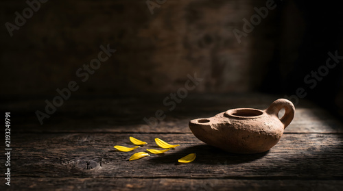Minimal still life of a single unlit clay oil lamp surrounded by flower petals on weathered dark wood, with dramatic natural sidelight and clean negative space