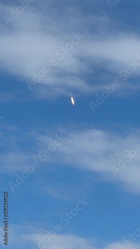 A rocket climbs through a bright blue sky with soft white clouds after liftoff. The glowing flame, vertical ascent, and open atmosphere create a dramatic aerospace launch scene.