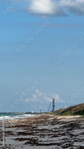 A rocket booster descends through the blue sky near a coastal launch pad. The beach, dunes, smoke, and landing flame show a dramatic spaceflight recovery scene.