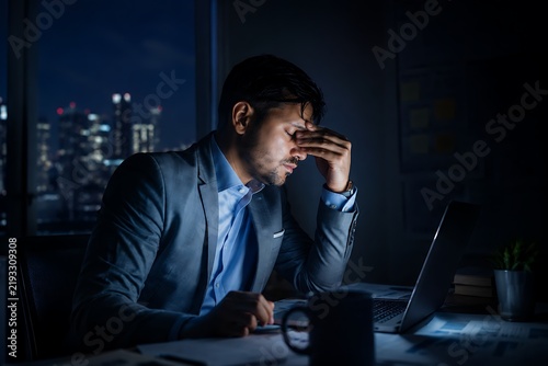A tired businessman working late at night in a dimly lit office. He is wearing a suit and tie, and has his hand on his forehead, appearing stressed or overwhelmed.