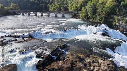 Aerial view of Rhine Falls in Switzerland with powerful cascades rushing between rocky formations, a Swiss flag, cliffside viewing paths and a historic railway bridge.