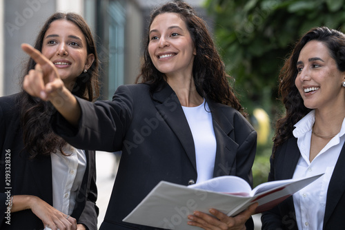 Foto A diverse team of three Hispanic businesswomen in dark suits, wide-eyed at what she sees, read an open report together outdoors with one pointing upward as the others look