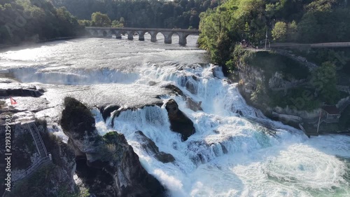 Aerial view of Rhine Falls in Switzerland with powerful cascading water, Schloss Laufen on the rocky hillside and a historic railway bridge surrounded by lush green forest.