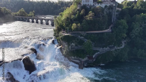 Aerial view of Rhine Falls in Switzerland with powerful cascading water, Schloss Laufen on the rocky hillside and a historic railway bridge surrounded by lush green forest.