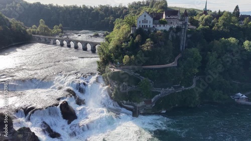 Aerial view of Rhine Falls in Switzerland with powerful cascading water, Schloss Laufen on the rocky hillside and a historic railway bridge surrounded by lush green forest.