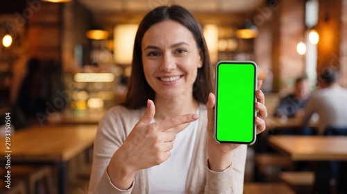 Woman showing green screen smartphone in cafe. Happy female holding mobile phone with chroma key display. Indoor restaurant photography for tech app.