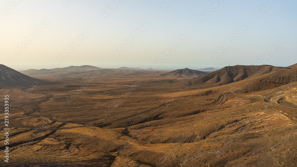 Fototapeta premium A sweeping view from Mirador de la Muda overlooks a vast arid valley and winding roads leading toward distant volcanic hills, Fuerteventura, Canary Islands, Spain.