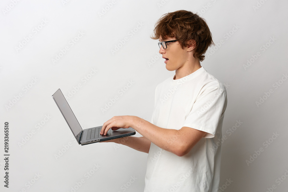 Obraz premium Young man in glasses and white t-shirt holds a laptop and reacts with surprise, mouth open. Side view on a gray background. Concept for news, technology or online sales.