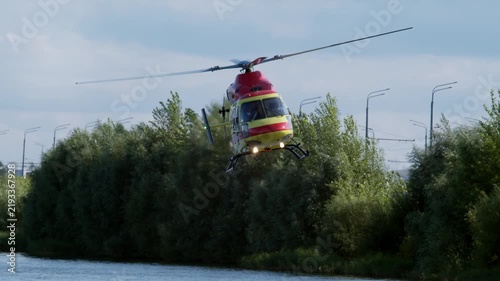 Red and yellow medical helicopter flies low over green trees on emergency dispatch mission. Air ambulance rushes to urgent call.