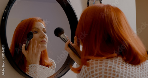 Фотография Redhead female applying foundation with makeup brush, carefully examining facial