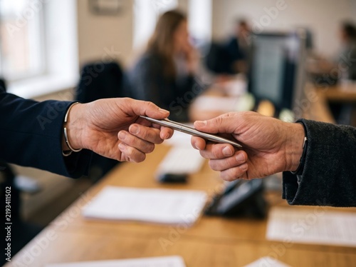 Close-up of two business professionals exchanging a silver pen above a wooden desk in a modern corporate office, ready to sign an important contract.