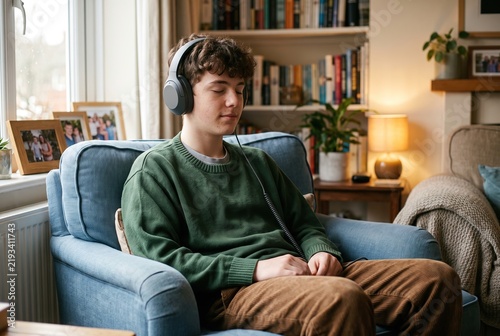A young person sits on a blue couch with headphones on, listening to music while relaxing at home. The room has bookshelves, a lamp, and family photos, creating a comfortable environment