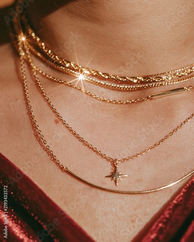 Close up fashion detail of layered gold necklaces on a model's neck, showcasing various styles and textures against a rich burgundy fabric background
