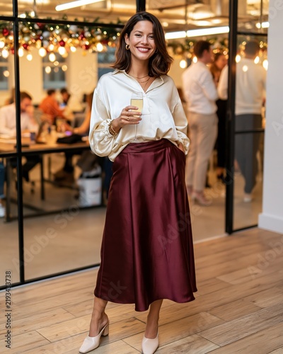 Fashion lifestyle editorial featuring a woman in a stylish cream blouse and burgundy satin skirt, holding a drink in a modern office setting with festive decorations