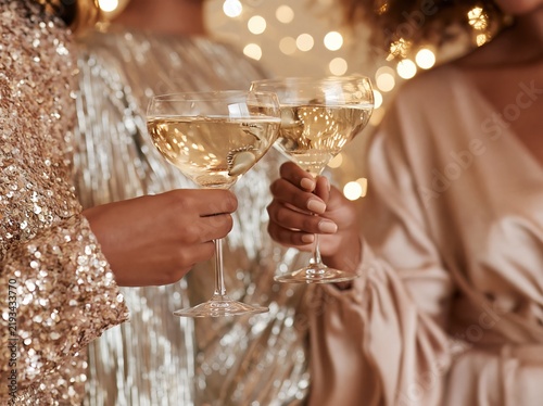 Three women holding champagne glasses in elegant dresses, celebrating with sparkling lights in the background, showcasing a festive atmosphere for a fashion lifestyle editorial