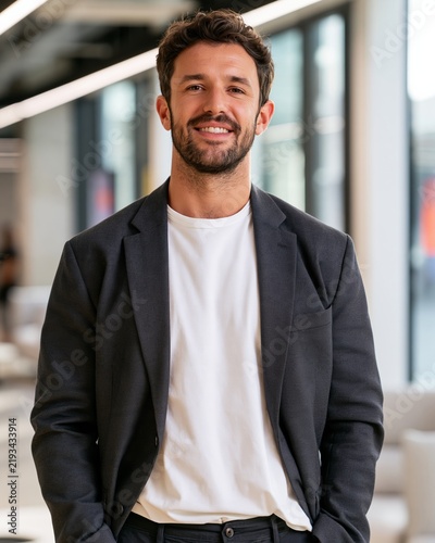 Fashion lifestyle portrait of a man in a black blazer and white t-shirt, smiling confidently in a modern office environment with large windows and contemporary furnishings