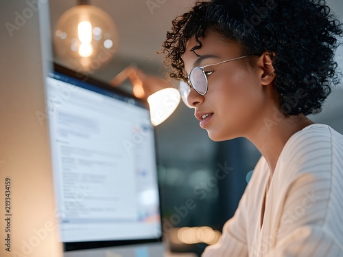 Professional African American woman with curly hair wearing glasses works on a computer in a modern office environment with warm lighting and blurred background elements