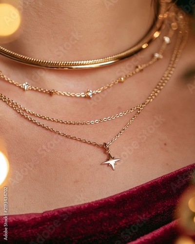 Close-up of layered gold necklaces featuring a star pendant on a womans neck, adorned with festive lights and a rich burgundy fabric background