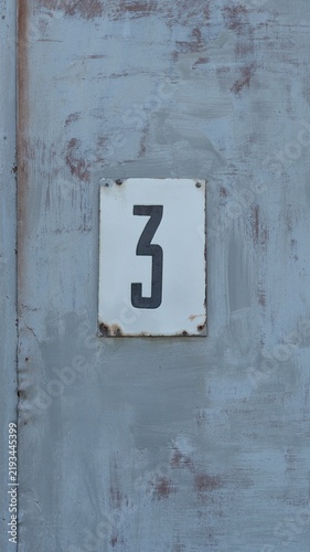 A white plate with the black number 3 mounted on a weathered grey painted metal gate. Close-up