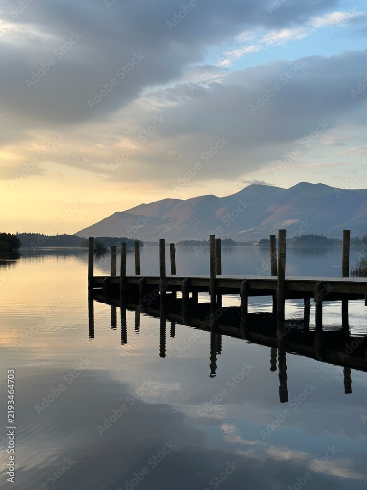 Fototapeta premium Wooden pier extending into a calm mountain lake at sunset, with dramatic clouds and beautiful reflections on the peaceful water.