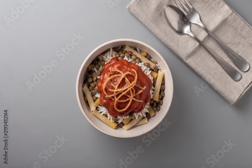 Koshary bowl filled with rice, pasta, lentils, chickpeas topped with fried onions, served on light grey surface. Concept of comfort food, vibrant colors enhance appetite.