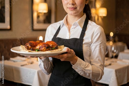 Waitress in dimly lit restaurant holding Musakhan on flatbread with chicken, onions, sumac, pine nuts. Concept of inviting ambiance with cozy booths and warm lighting.