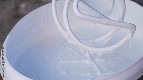 Universal mixer tool spins in bucket of white paint, achieving required consistency for later application on surfaces. This step is essential for even distribution during construction or repair work