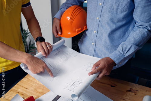 Architects and engineers discussing building blueprint at desk with model house, safety helmet, and tools, focusing on teamwork, planning, and precision in construction project.
