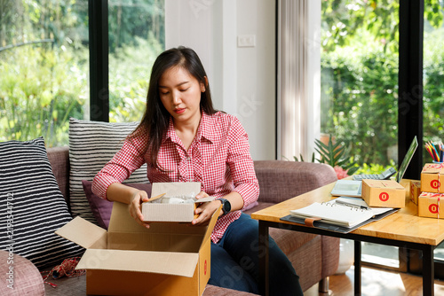 Young female entrepreneur packing a small parcel into a larger cardboard box on a sofa. Online merchant working from home preparing customer orders for shipping.