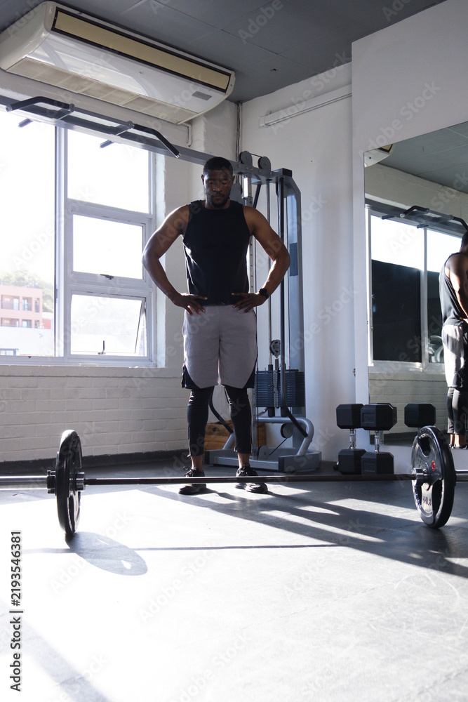 Fototapeta premium African man standing hands on hips facing barbell in gym wearing black sleeveless top, gray shorts