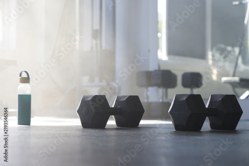 Pair of black hex dumbbells are resting on dark rubber gym floor near green water bottle