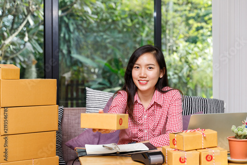 Smiling young Asian woman online seller holding parcel box at home office desk with cardboard packages. E-commerce SME small business, order fulfillment, and shipping concept.